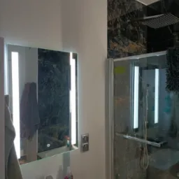 Nowoczesna łazienka z czarnymi płytkami imitującymi marmur, podświetlane lustro nad umywalką, kabina prysznicowa typu walk-in z deszczownicą.