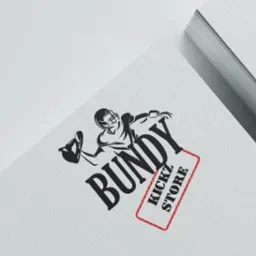 Czarno-białe logo przedstawiające gracza futbolu amerykańskiego rzucającego butem, obok nazwy 'BUNDY KICKZ STORE' w czerwonej ramce, umieszczone na białym papierze z plikiem kartek w tle.