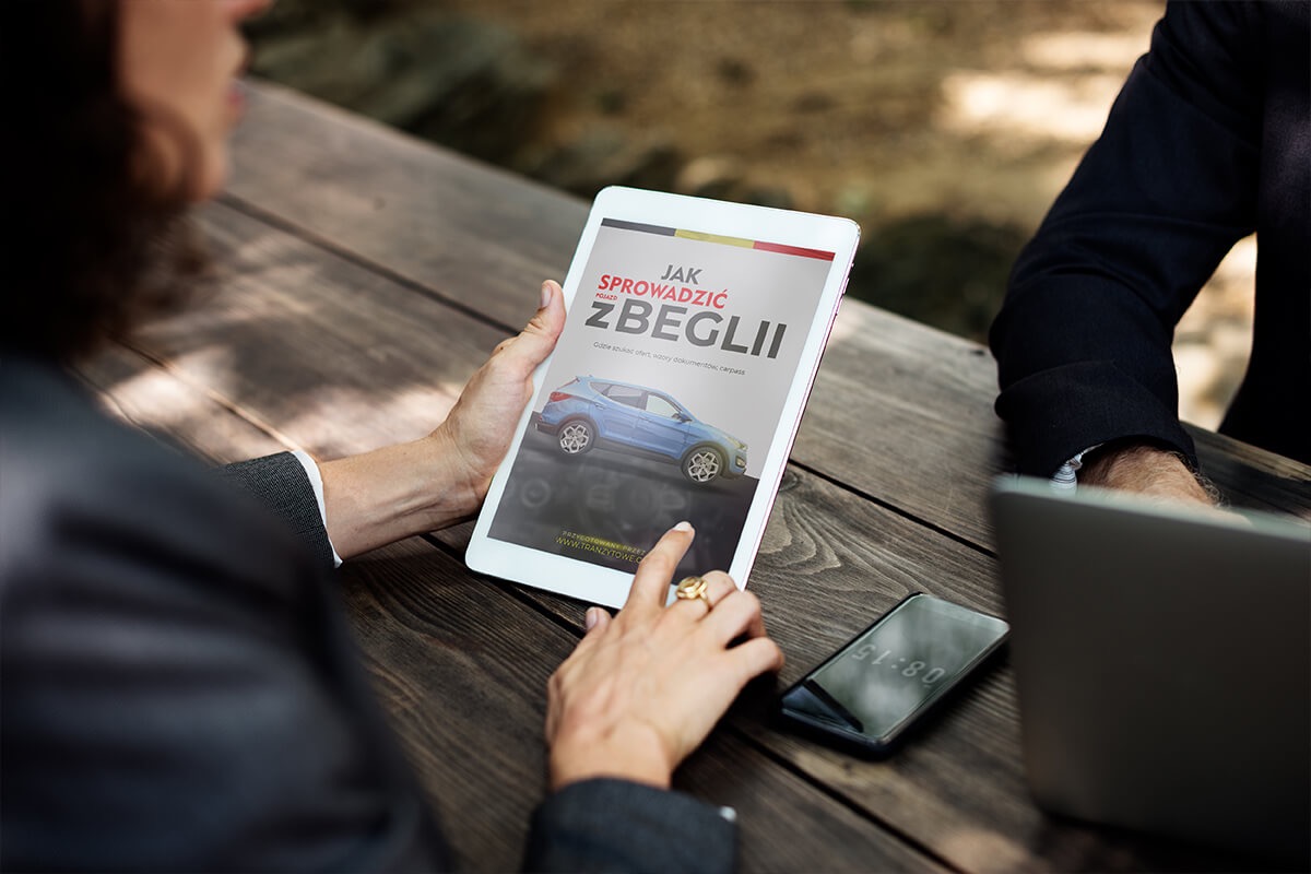 Osoba przegląda na tablecie okładkę e-booka o sprowadzaniu samochodów z zagranicy, na drewnianym stole leży smartfon i laptop.