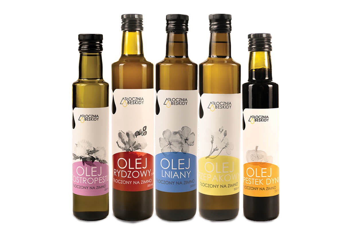 Pięć butelek oleju tłoczonego na zimno, każda z inną etykietą i grafiką kwiatową, ustawione obok siebie na białym tle. Oleje różnią się kolorem, od jasnożółtego po ciemnobrązowy.
