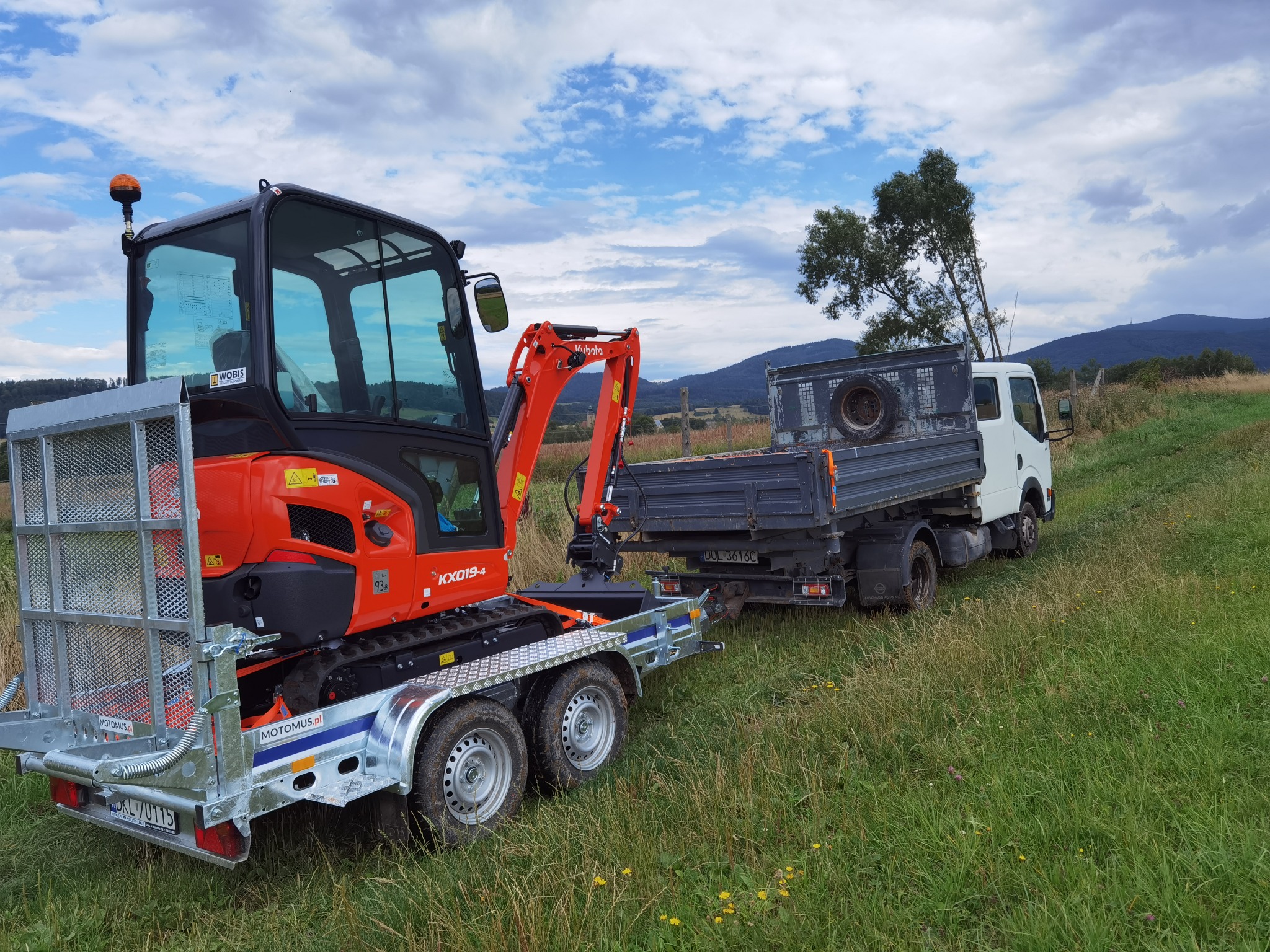 Pomarańczowa mini koparka Kubota KX019-4 na przyczepie, ciągnięta przez białą ciężarówkę na tle zielonej łąki i gór.