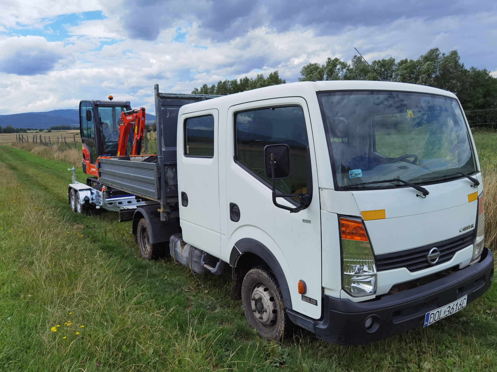 Biała ciężarówka Nissan Cabstar z przyczepą, na której przewożony jest pomarańczowy minikoparka Kubota, zaparkowana na trawiastym poboczu drogi polnej z widokiem na odległe góry i pochmurne niebo.