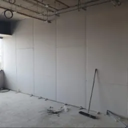 Montaż płyt karton gips na ścianie w surowym wnętrzu, widoczne kable, szczotka i elementy mocujące na podłodze, duże okno z widokiem na budynek.