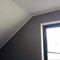 Pomieszczenie po malowaniu ścian na szaro, widoczne okno z czarną ramą i białe wykończenie sufitu na poddaszu.