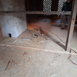 Adrian Sosna firma ogólnobudowlana - Płyty OSB na poddaszu w trakcie układania, widoczne narzędzia, poziomica i wiertarka, fragment ściany z ubytkiem tynku, osoba w tle.