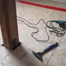 Montaż podłogi z płyt OSB na poddaszu, widoczny drewniany słup, poziomica, wiertarka i młotek dekarski.