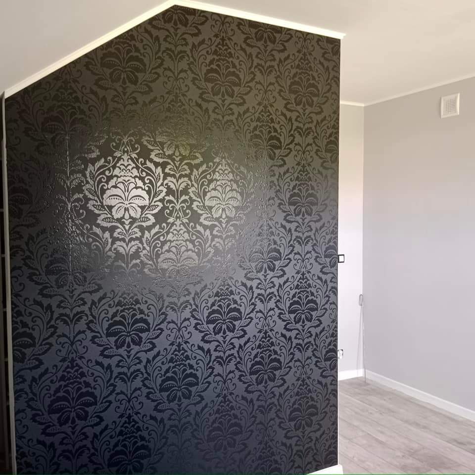 Ściana pokryta ciemną tapetą w ornamenty roślinne, z widocznym przejściem do jasnoszarego pomieszczenia z drewnopodobną podłogą.