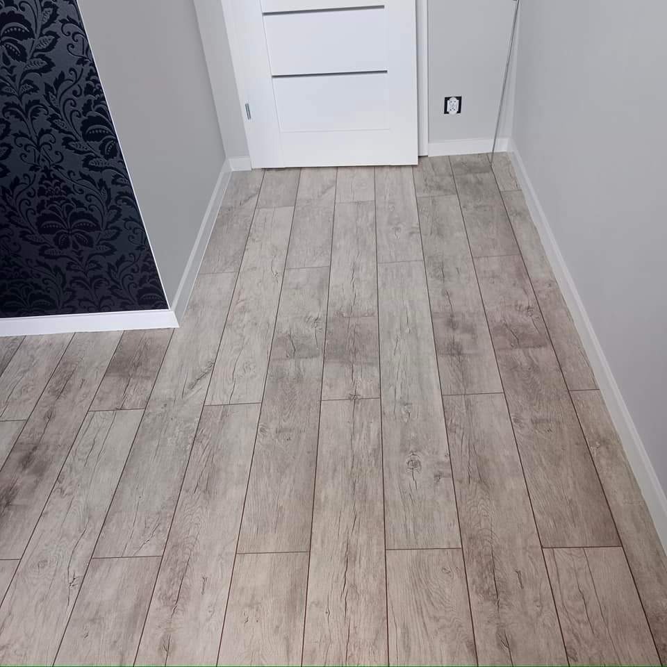 Ułożony parkiet laminowany w odcieniach szarości z widoczną strukturą drewna, ściana pomalowana na szaro z dekoracyjną tapetą w kwiaty, białe listwy przypodłogowe i białe drzwi w tle.