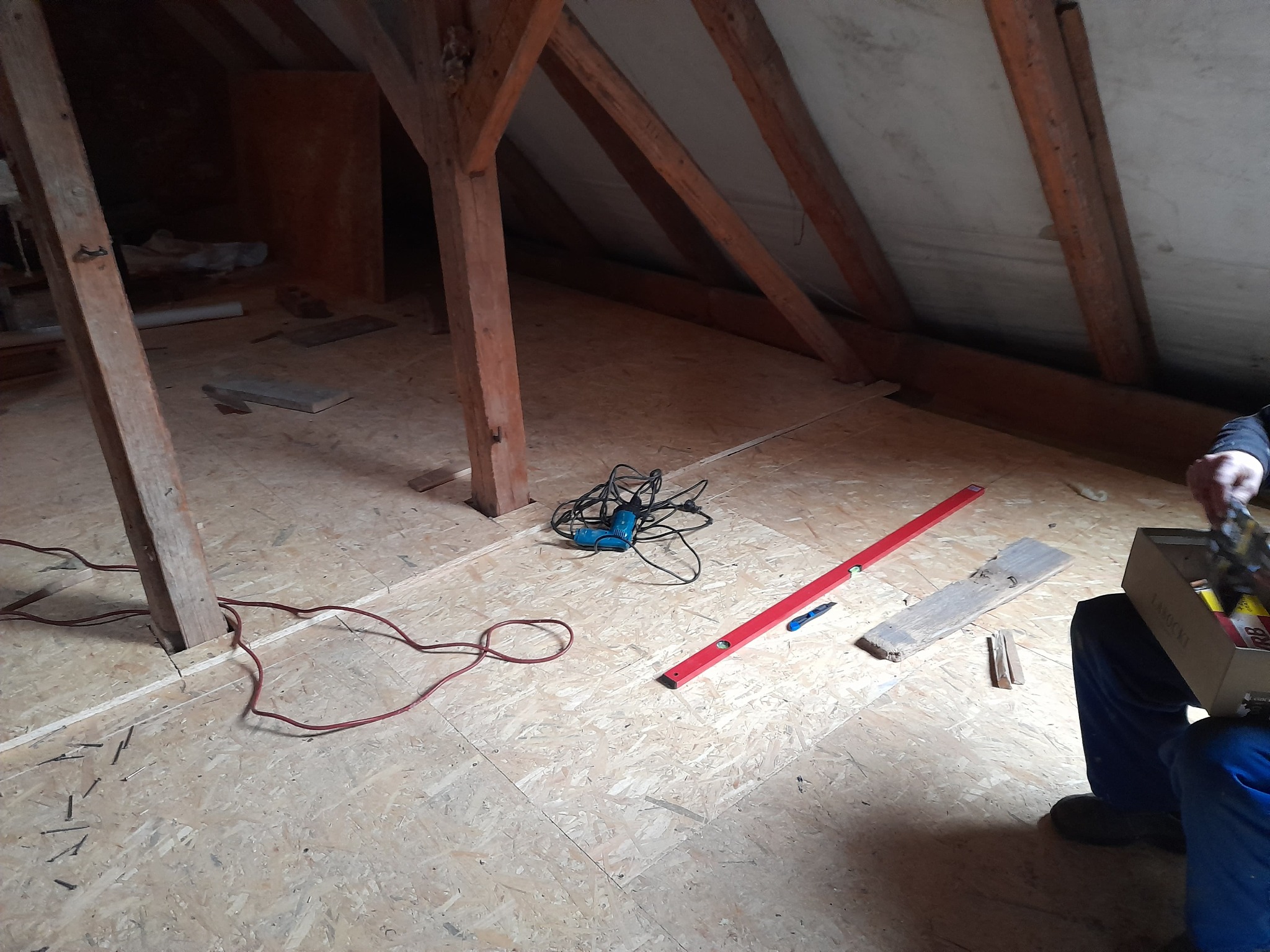 Poddasze w trakcie prac adaptacyjnych: surowa drewniana konstrukcja, płyty OSB na podłodze, poziomica, wiertarka i narzędzia przygotowane do dalszych prac wykończeniowych.