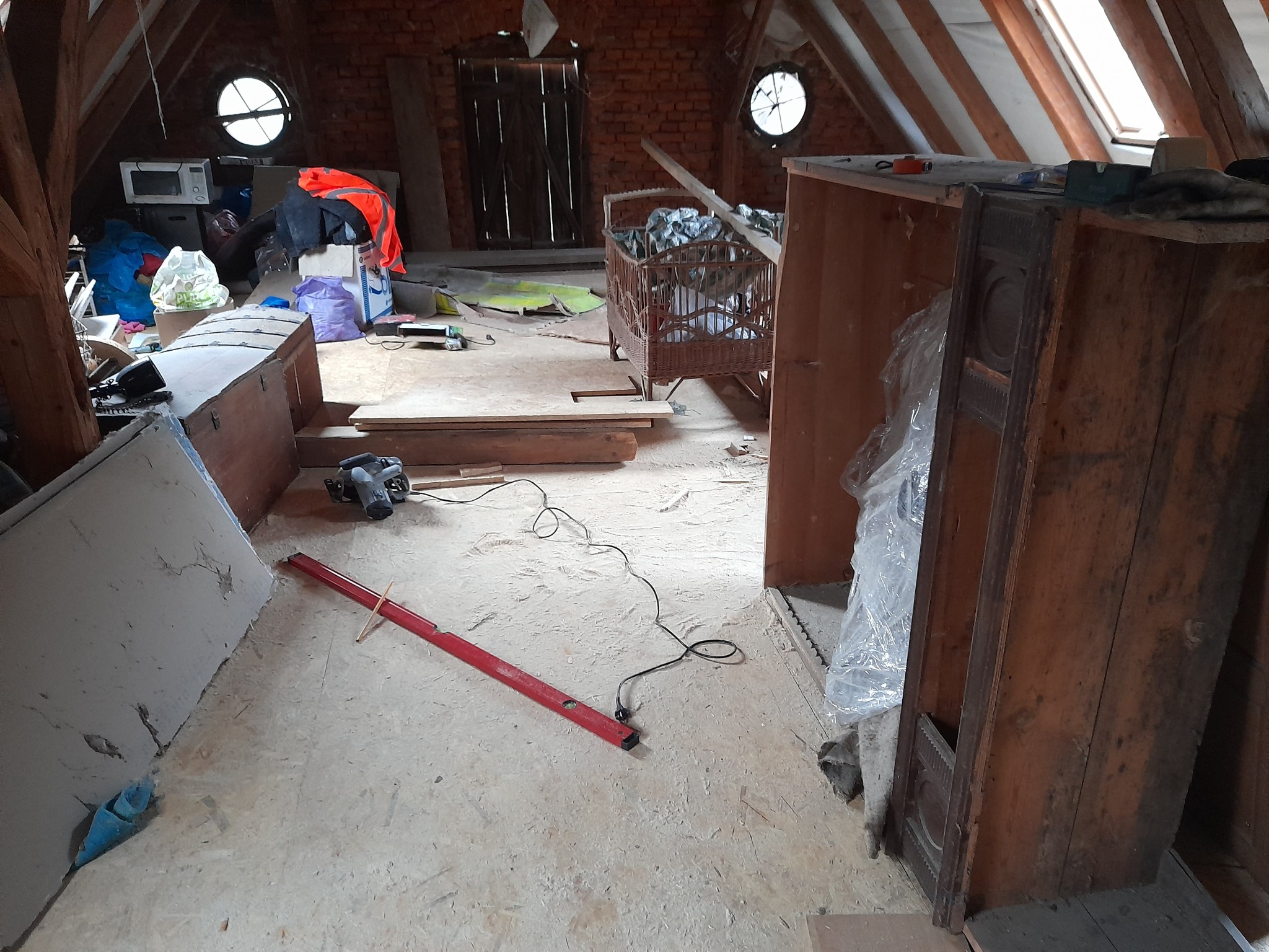 Poddasze w trakcie adaptacji: widoczne krokwie, okna połaciowe, narzędzia, poziomica, płyta gipsowa, meble i bałagan.