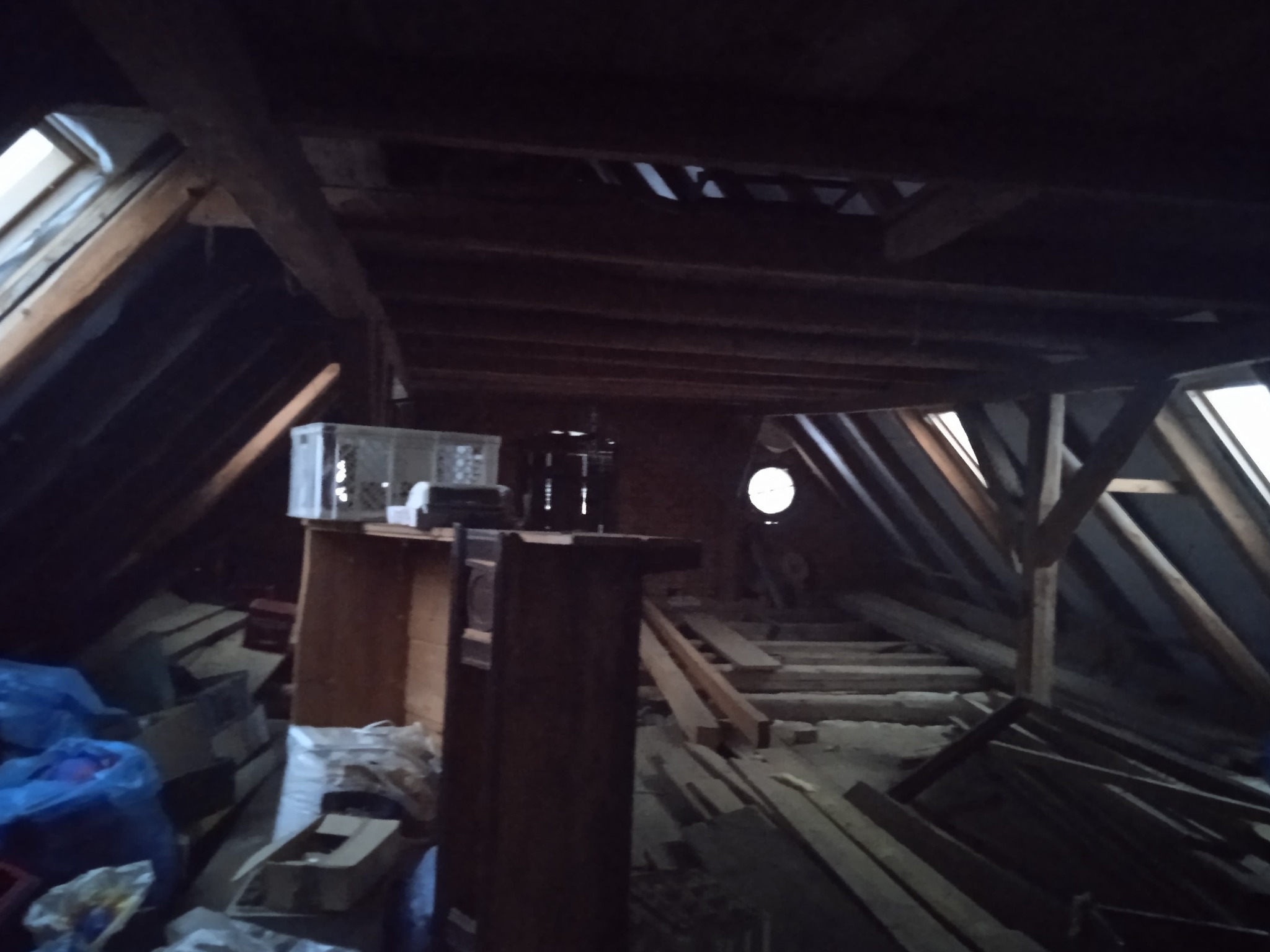Słabo oświetlone poddasze z widoczną konstrukcją dachu, fragmentami desek i oknem dachowym, w tle okrągłe okno, częściowo zagracone materiałami i przedmiotami.