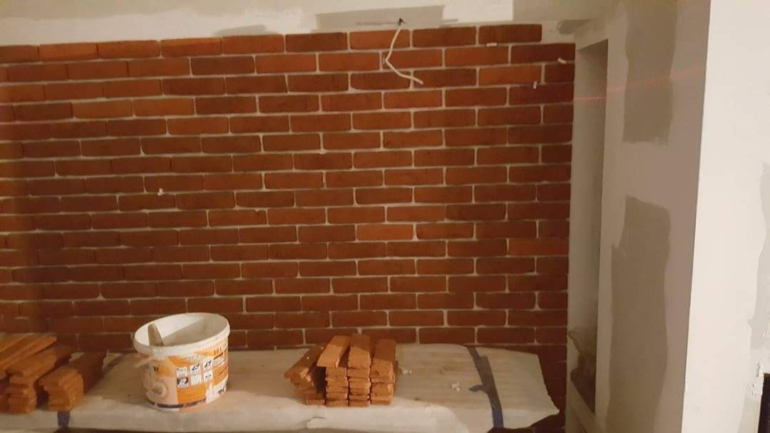 Ściana wyłożona cegiełkami, widoczny pojemnik z klejem oraz stos cegiełek na podłożu zabezpieczonym folią malarską, obok fragment ściany przygotowanej do malowania.