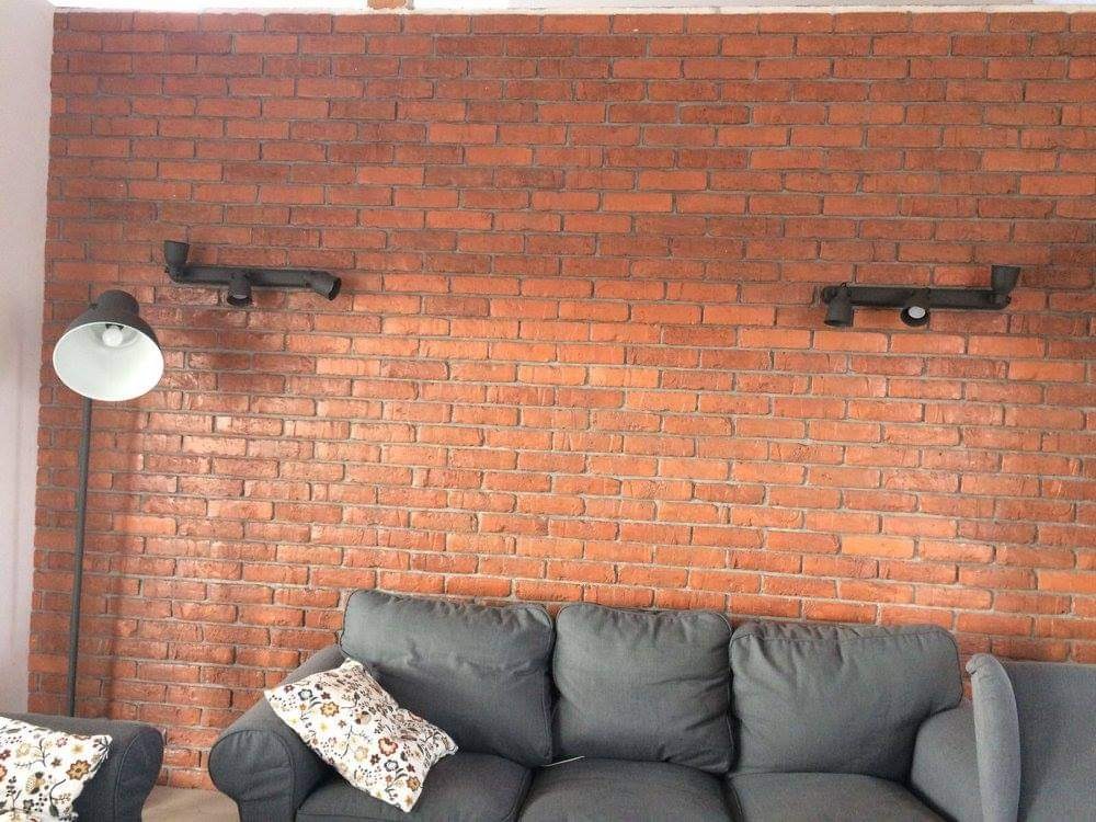 Czerwona ściana z cegły z dwoma czarnymi lampami rurowymi, szara sofa z poduszkami w kwiaty i lampa podłogowa z białym kloszem.