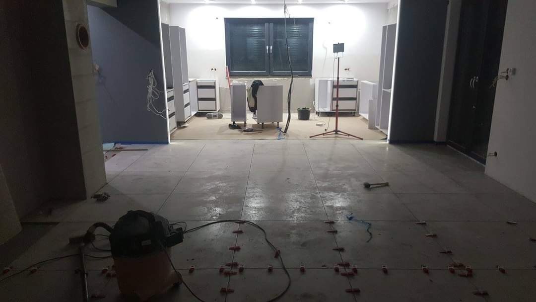 Szare płytki podłogowe w trakcie układania, widoczne krzyżyki dystansowe, odkurzacz przemysłowy i meble kuchenne w tle.