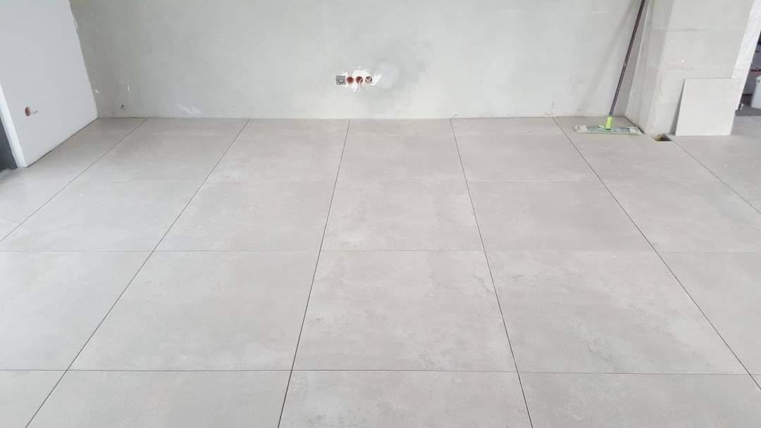 Nowo ułożona, jasnoszara podłoga z płytek ceramicznych w surowym wnętrzu, widoczne gniazdka elektryczne bez osprzętu i mop do czyszczenia podłogi w rogu pomieszczenia.