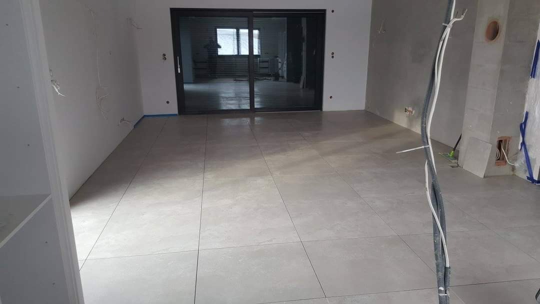 Wnętrze w trakcie prac wykończeniowych: ułożona jasnoszara podłoga z płytek, ściany przygotowane pod malowanie, widoczne wystające kable i rury instalacyjne, duże przesuwne okno tarasowe w tle.