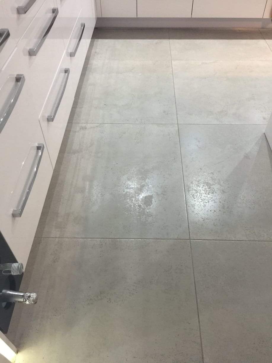 Szare, matowe płytki podłogowe w kuchni, widoczne fugi i fragment szafek kuchennych z białymi frontami i srebrnymi uchwytami.