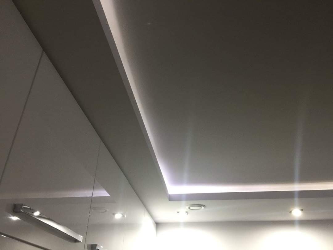 Sufit podwieszany z oświetleniem LED wzdłuż krawędzi, widoczne również punkty oświetleniowe i fragment ściany z szafką z połyskiem.