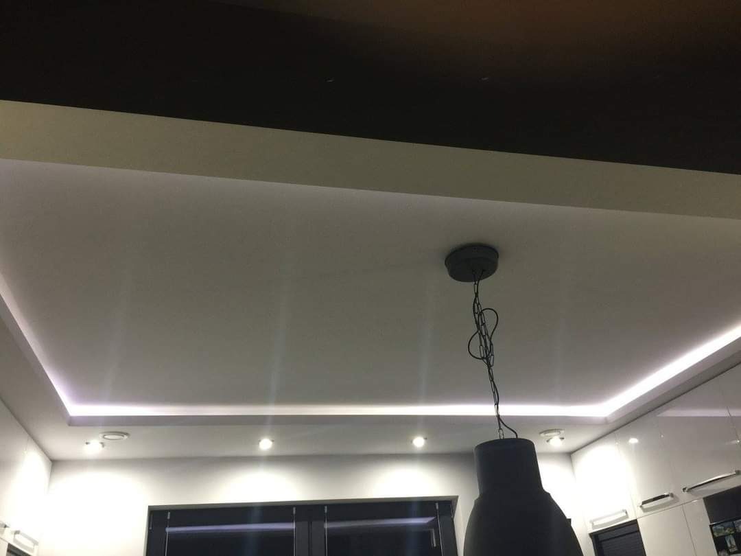 Nowoczesny sufit podwieszany z oświetleniem LED w kształcie prostokąta, widoczna czarna lampa wisząca na łańcuchu, wnętrze kuchni z białymi szafkami w tle.