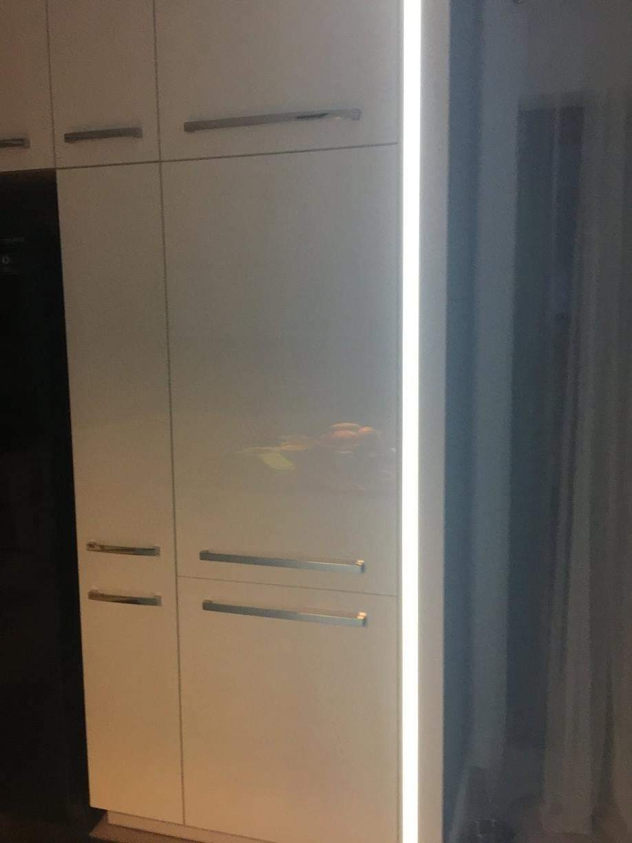 Biała zabudowa meblowa z połyskiem, podświetlona pionową listwą LED, z poziomymi, metalowymi uchwytami.