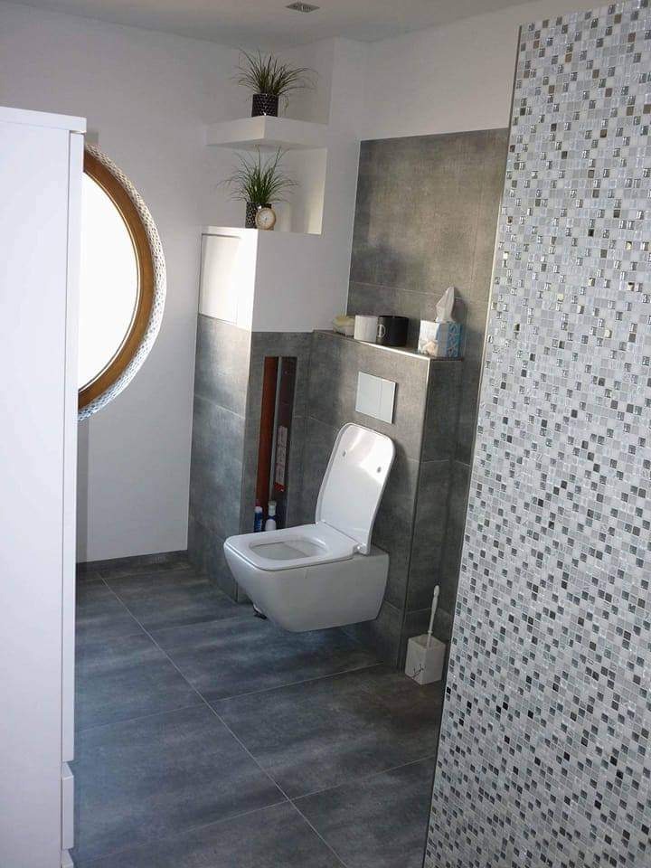 Nowoczesna łazienka z wiszącą toaletą, szarą płytką podłogową i ścianą wyłożoną mozaiką w odcieniach szarości i bieli; na półkach dekoracyjne rośliny w doniczkach.