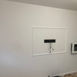 Karol Matuszewski - Biała ściana po malowaniu z dekoracyjną ramą, uchwytem do telewizora z kablami oraz zamontowanym biokominkiem.