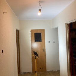 Karol Matuszewski - Korytarz w trakcie odświeżania: ściany częściowo pomalowane na jasny kolor, widoczna lampa budowlana na statywie, stara terakota na podłodze, otwory po gniazdkach elektrycznych w ścianie.