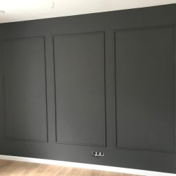 Karol Matuszewski - Ściana pomalowana na szaro z dekoracyjnymi listwami tworzącymi trzy prostokątne panele, widoczne gniazdka elektryczne przy podłodze i fragment drewnianej podłogi.