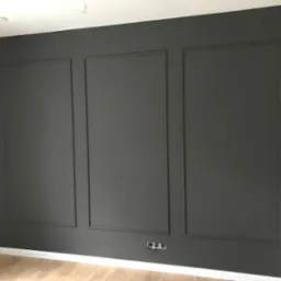 Ściana pomalowana na szaro z dekoracyjnymi listwami tworzącymi trzy prostokątne panele, widoczne gniazdka elektryczne przy podłodze i fragment drewnianej podłogi.