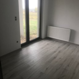 Karol Matuszewski - Pokój po remoncie z nowymi panelami podłogowymi, grzejnikiem na ścianie i widokiem na ogród przez duże okno balkonowe; widoczne puszki elektryczne bez osprzętu.
