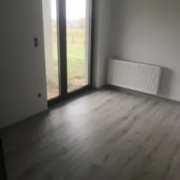 Pokój po remoncie z nowymi panelami podłogowymi, grzejnikiem na ścianie i widokiem na ogród przez duże okno balkonowe; widoczne puszki elektryczne bez osprzętu.