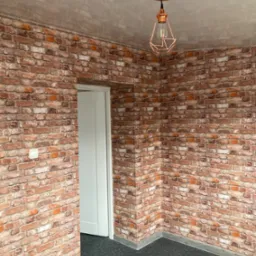 Pokój z tapetą imitującą czerwoną cegłę, szarą podłogą i miedzianą lampą wiszącą. Białe drzwi w tle. Widoczny fragment ściany i sufitu.