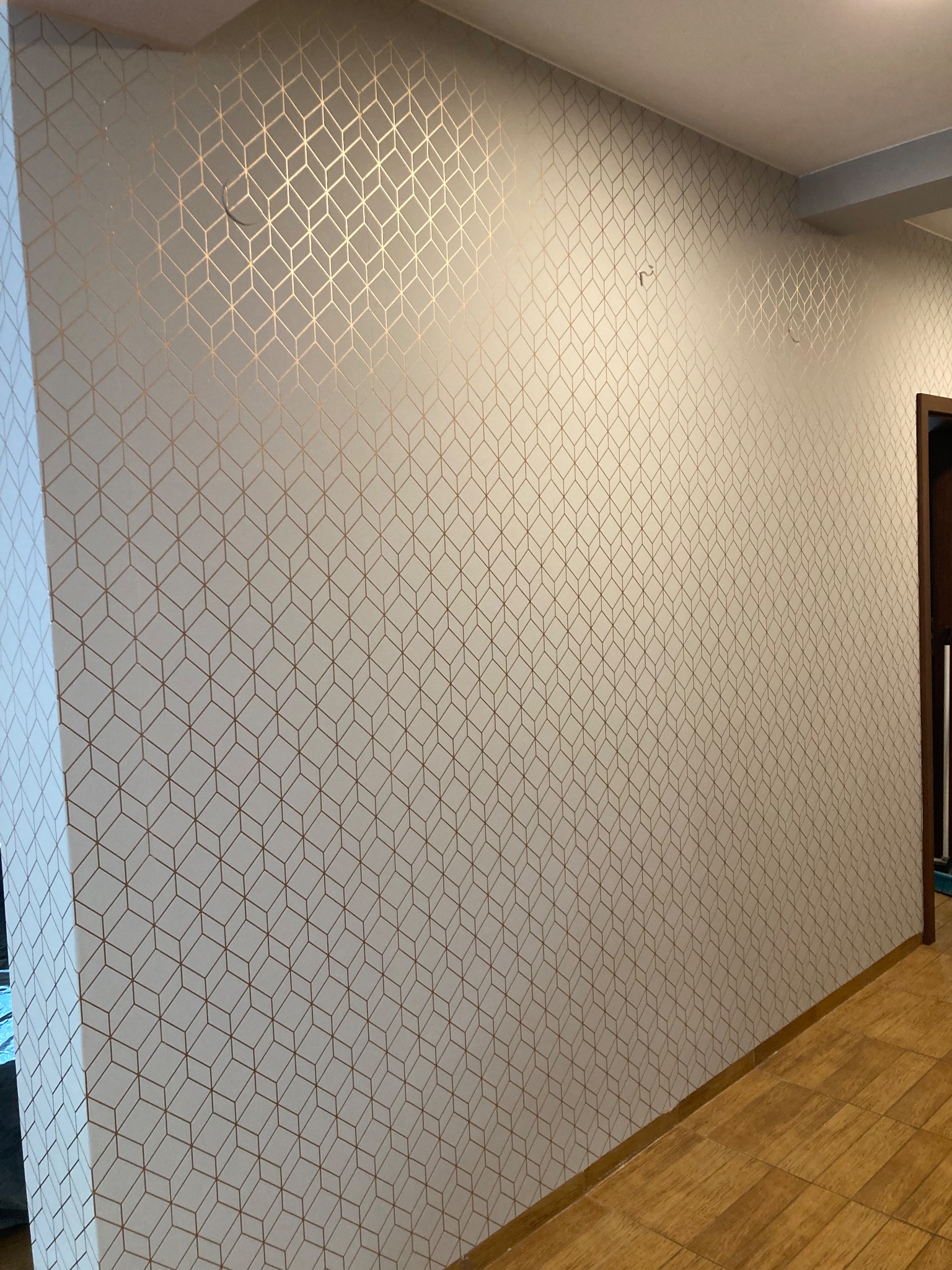 Ściana w korytarzu pokryta białą tapetą z geometrycznym wzorem złotych rombów, widoczne fragmenty drewnianej podłogi i futryny drzwi