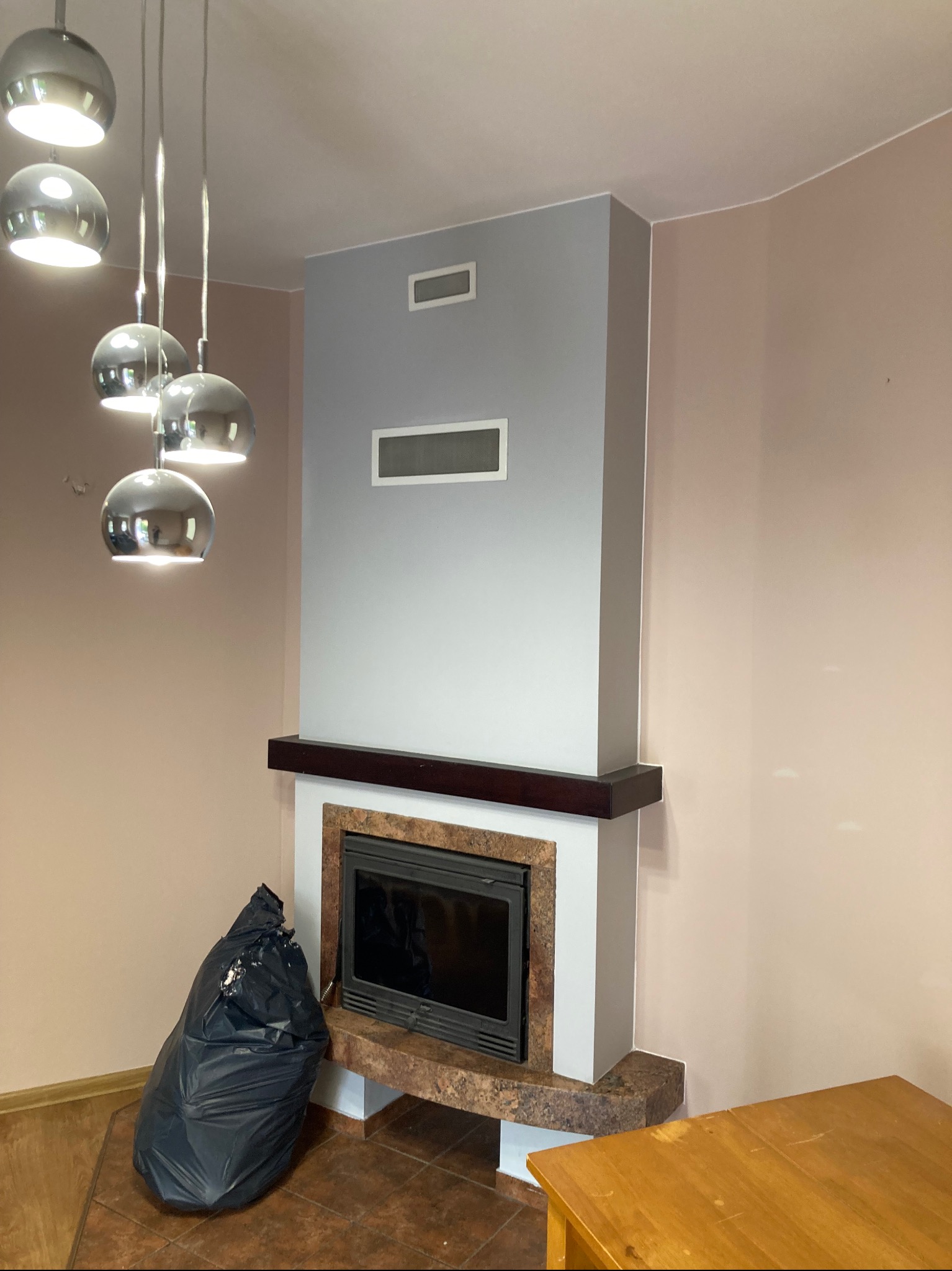 Pomalowany na szaro kominek z marmurowym obramowaniem, obok stoi worek na śmieci, ściany w odcieniach beżu i szarości, sufit oświetlają nowoczesne lampy kuliste.