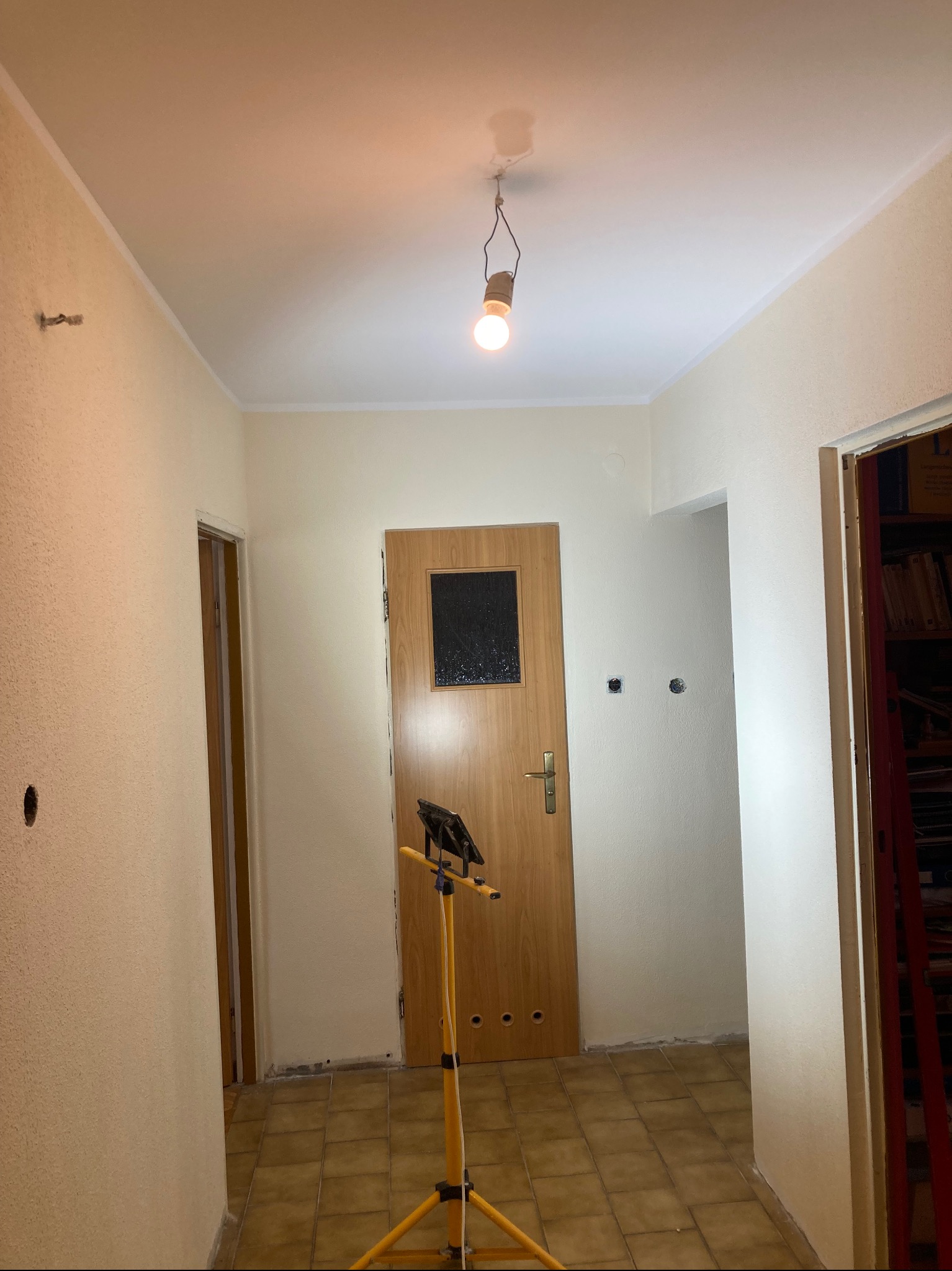 Korytarz w trakcie odświeżania: ściany częściowo pomalowane na jasny kolor, widoczna lampa budowlana na statywie, stara terakota na podłodze, otwory po gniazdkach elektrycznych w ścianie.