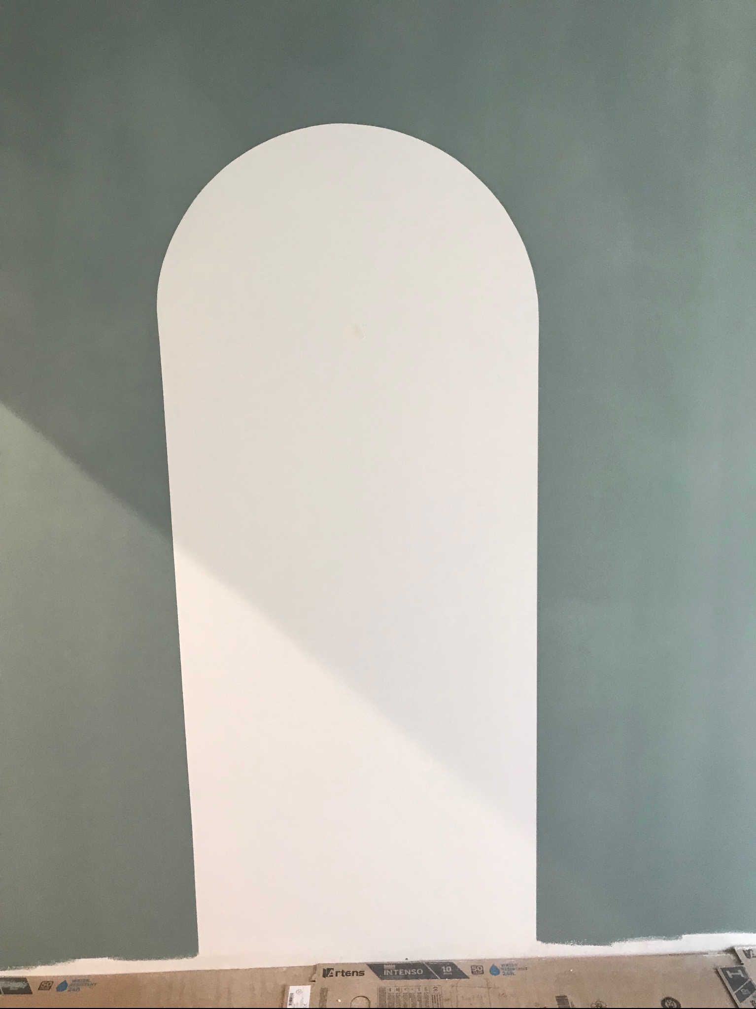 Świeżo pomalowana ściana w odcieniach zieleni i bieli, z widocznym odcięciem łukowym i kartonami na podłodze.