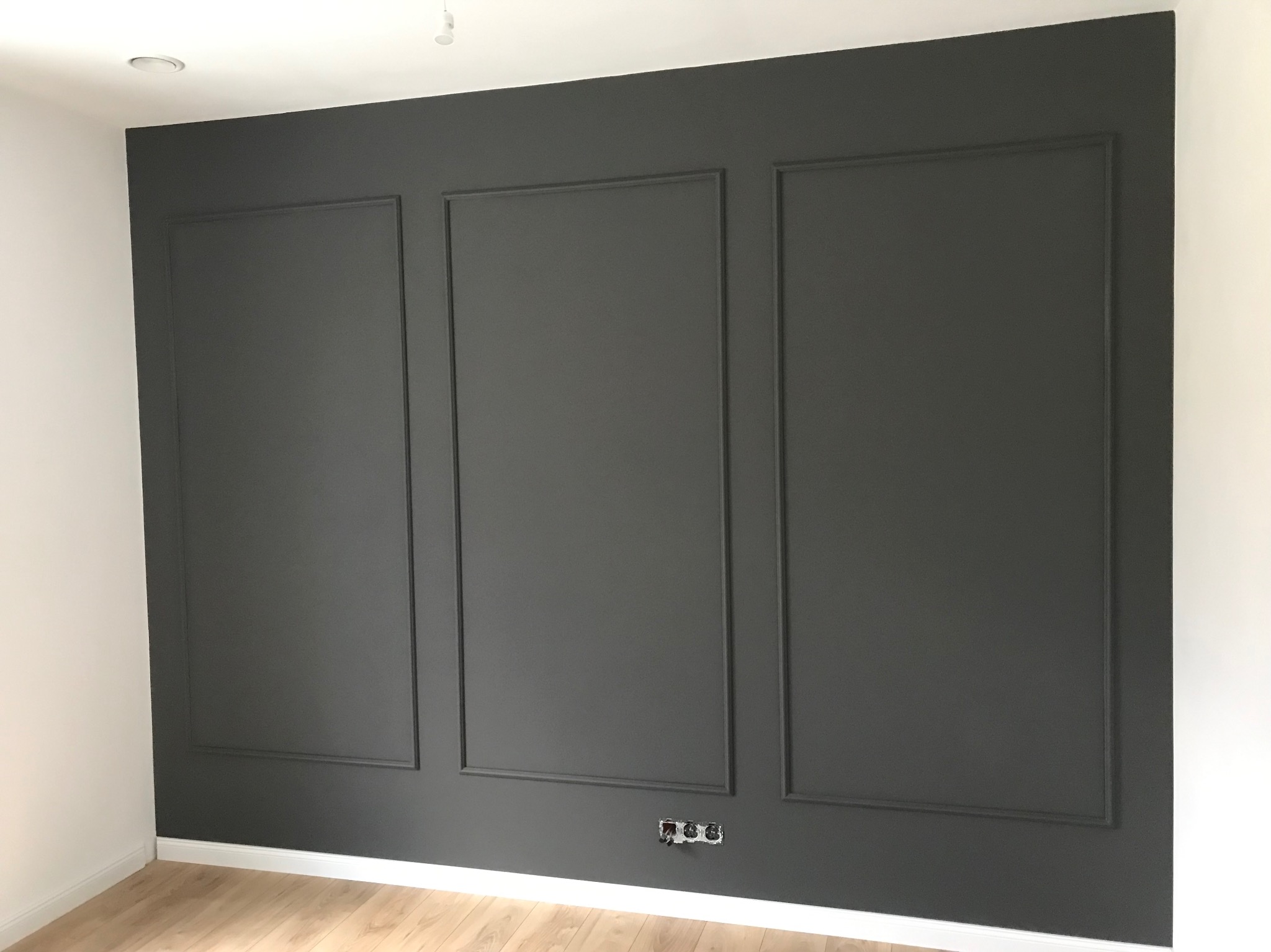 Ściana pomalowana na szaro z dekoracyjnymi listwami tworzącymi trzy prostokątne panele, widoczne gniazdka elektryczne przy podłodze i fragment drewnianej podłogi.