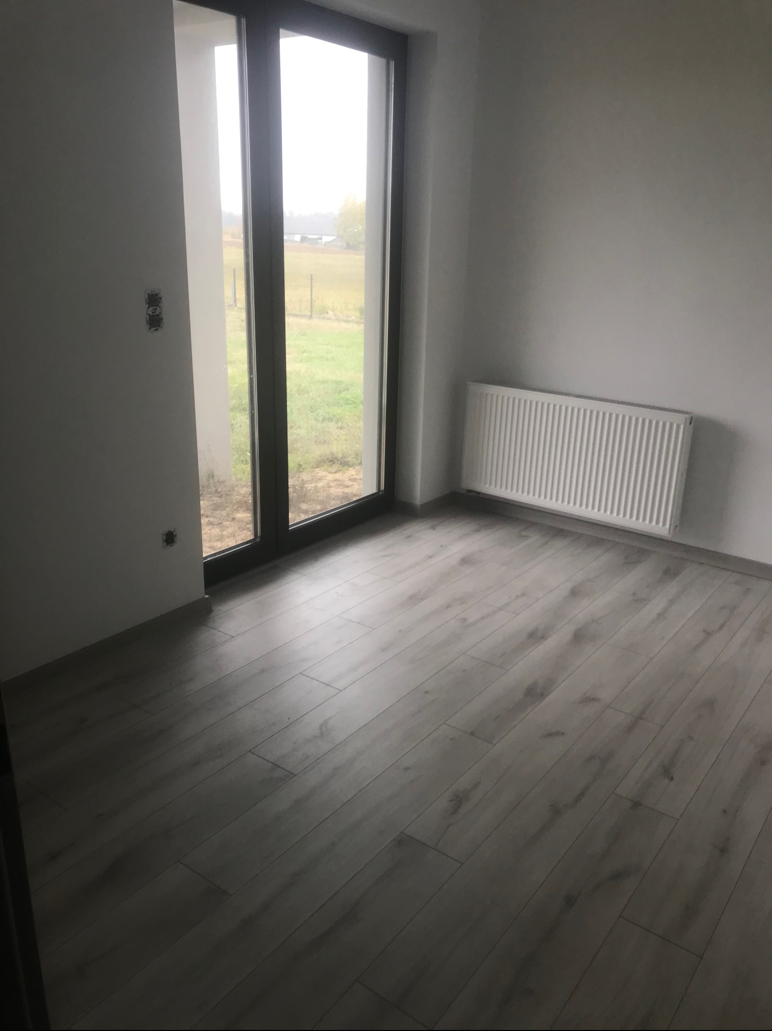 Pokój po remoncie z nowymi panelami podłogowymi, grzejnikiem na ścianie i widokiem na ogród przez duże okno balkonowe; widoczne puszki elektryczne bez osprzętu.