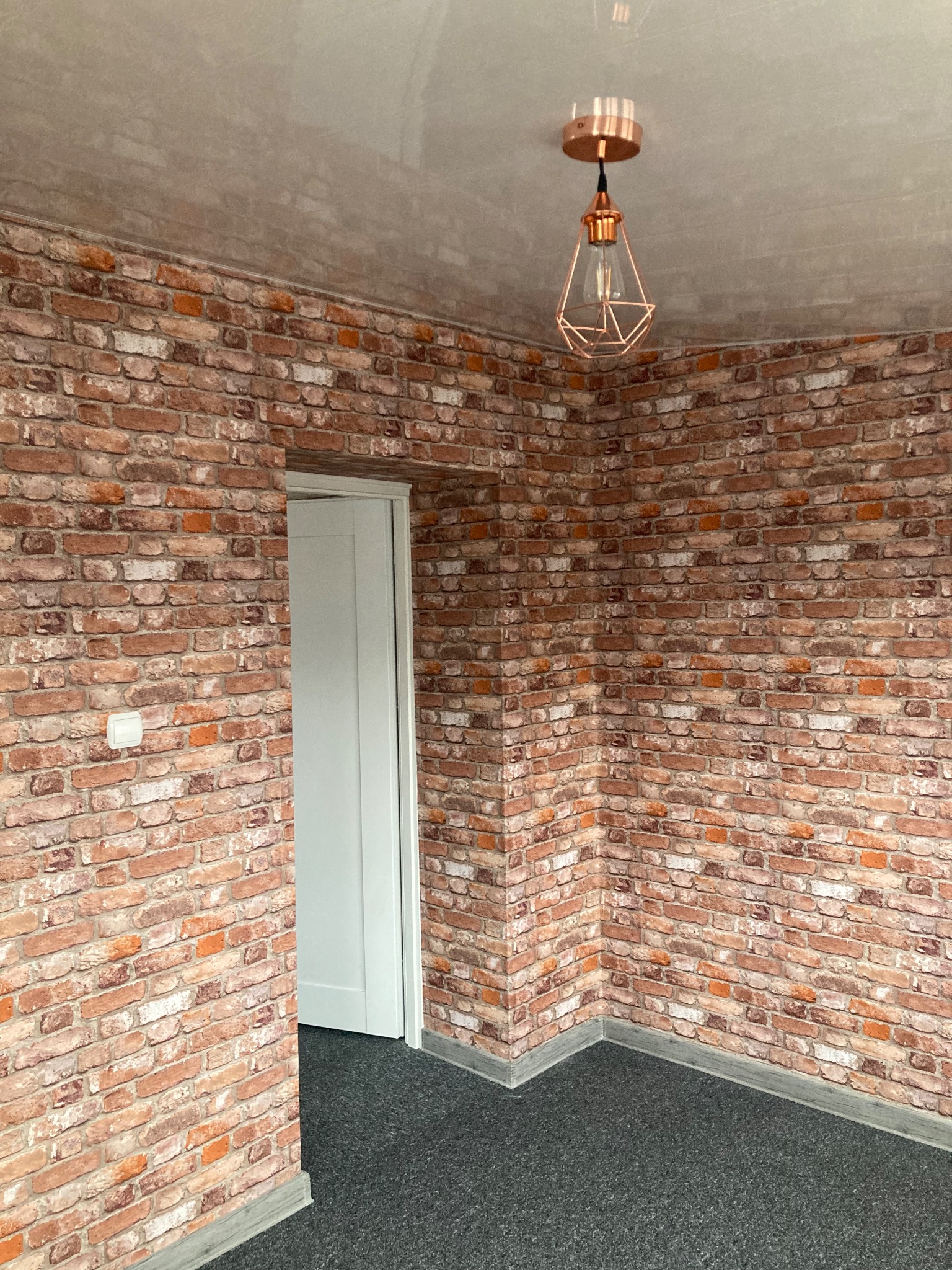 Pokój z tapetą imitującą czerwoną cegłę, szarą podłogą i miedzianą lampą wiszącą. Białe drzwi w tle. Widoczny fragment ściany i sufitu.