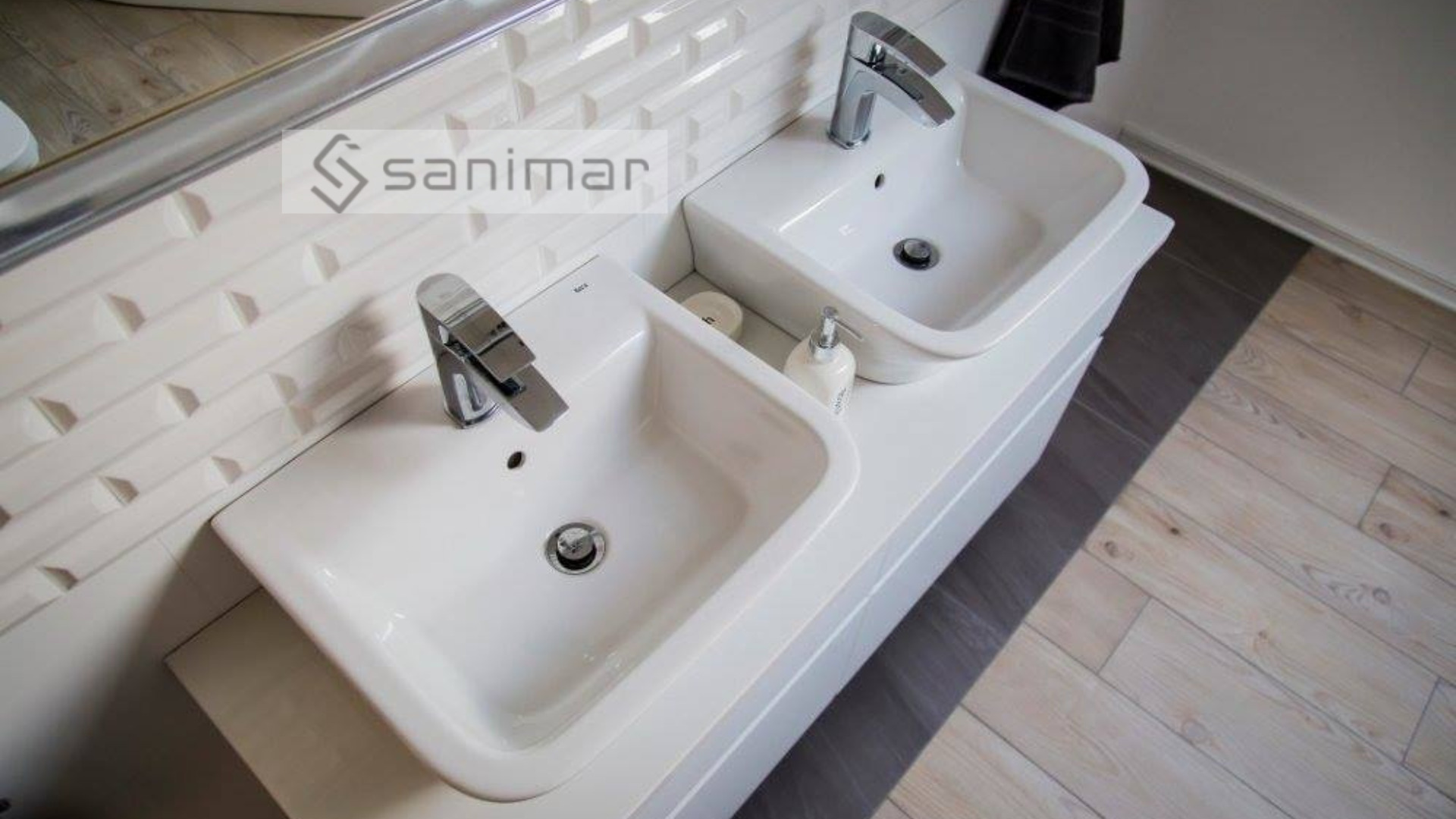 Dwie białe, prostokątne umywalki nablatowe z chromowanymi bateriami, ustawione obok siebie na białej szafce. Na blacie między umywalkami mydło w płynie i mydło w kostce. Tło to białe, trójwymiarowe...