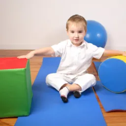 Chłopiec w białym ubraniu siedzi na niebieskiej macie z kolorowymi rekwizytami (kostka, wałek, piłka) podczas zajęć terapeutycznych.