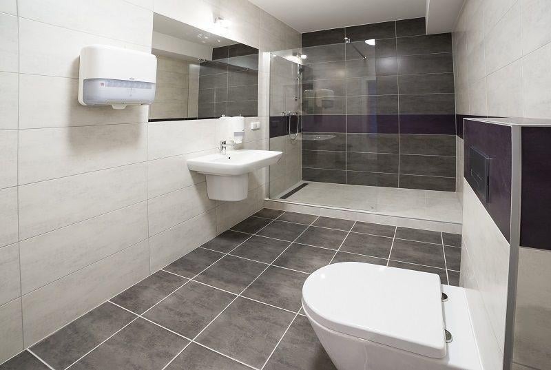 Nowoczesna łazienka z kabiną prysznicową typu walk-in, umywalką i toaletą, wykończona szarymi i białymi płytkami ceramicznymi z dyspenserem na ręczniki papierowe.