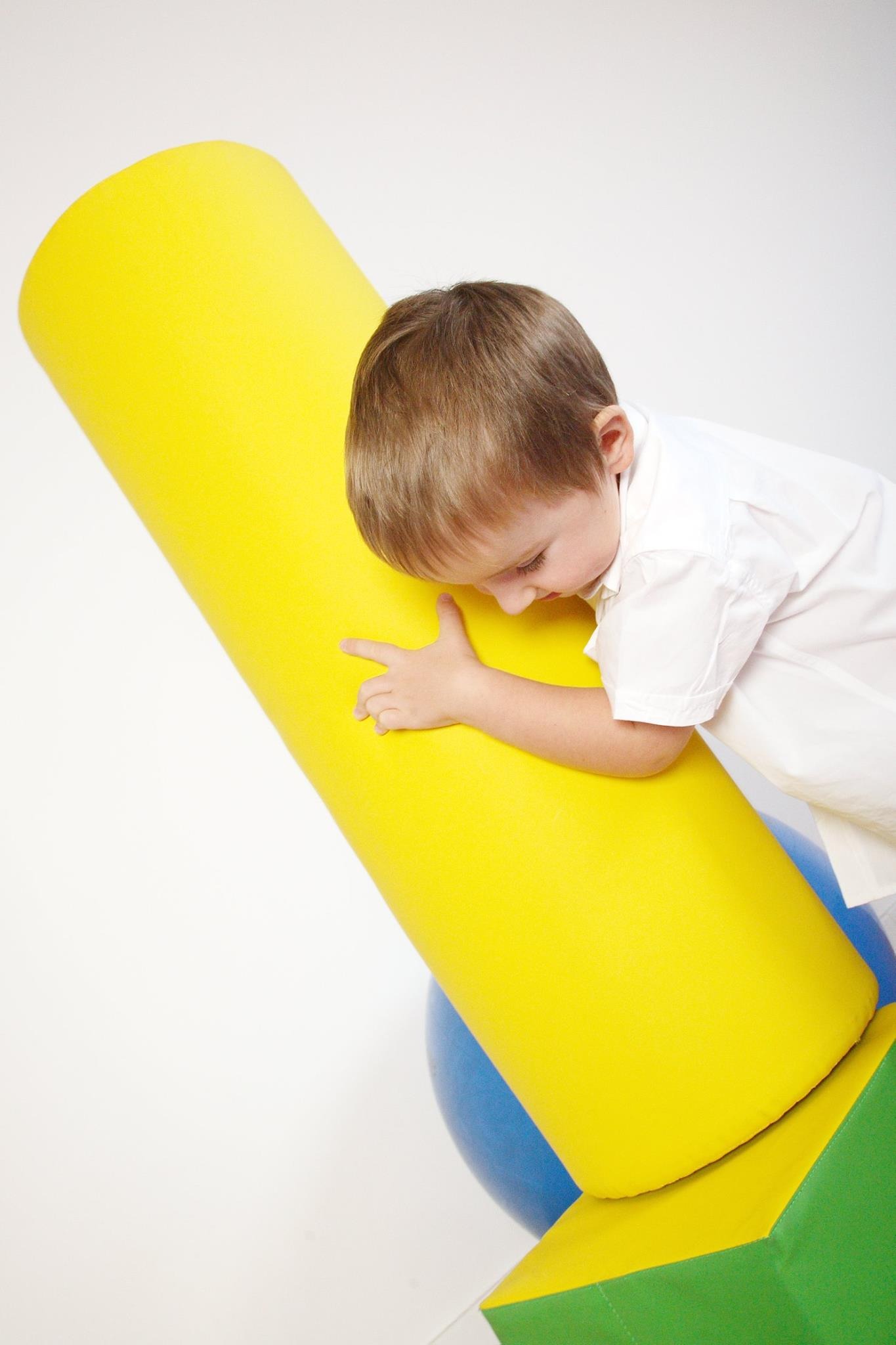Mały chłopiec w białej koszuli przytula się do dużego, żółtego, piankowego wałka, elementu wyposażenia sali do ćwiczeń lub rehabilitacji dla dzieci.