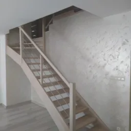 Jasne drewniane schody z metalowymi poręczami w nowoczesnym wnętrzu z dekoracyjną ścianą w odcieniach beżu i szarości.