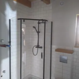 Nowoczesna kabina prysznicowa z czarnymi profilami i białą armaturą na tle białych, prostokątnych płytek, uzupełniona drewnianymi elementami dekoracyjnymi pod sufitem.