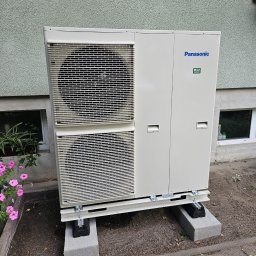 Zielony Program Ekologiczny sp. z o.o. - Zewnętrzna jednostka klimatyzacyjna Panasonic R32 na betonowych podstawach, z kwiatami w tle, ustawiona przy ścianie budynku.