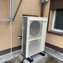 Zielony Program Ekologiczny sp. z o.o. - Zewnętrzna jednostka klimatyzacyjna Panasonic, zamontowana na betonowych podstawach przy ścianie budynku z brązową stolarką okienną i rynną odpływową, widoczne elementy instalacji.