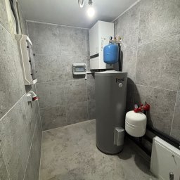 Zielony Program Ekologiczny sp. z o.o. - Wnętrze kotłowni z szarymi płytkami, centralnym zbiornikiem Ferroli, białym kotłem wiszącym i niebieskim zbiornikiem przeponowym. Widoczne panele elektryczne na ścianie.