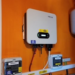 Zielony Program Ekologiczny sp. z o.o. - Biały falownik SOFAR zamontowany na pomarańczowej ścianie, pod nim skrzynki rozdzielcze PV-AC i PV-DC, widoczne rury instalacji.