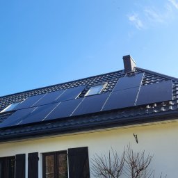 Zielony Program Ekologiczny sp. z o.o. - Czarny dach z panelami słonecznymi i oknami dachowymi na białym domu z ciemnymi okiennicami, widok z dołu na tle błękitnego nieba.